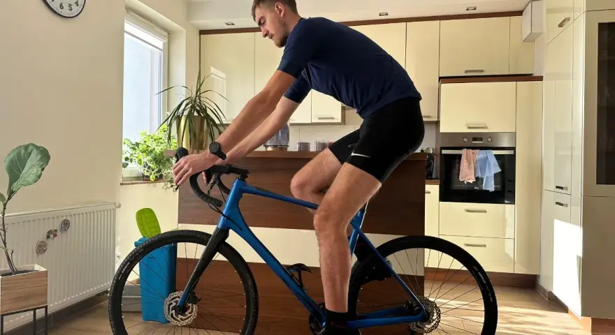 Mężczyzna w pozycji na rowerze gravel, trenuje w domu. W tle kuchnia i zegar.