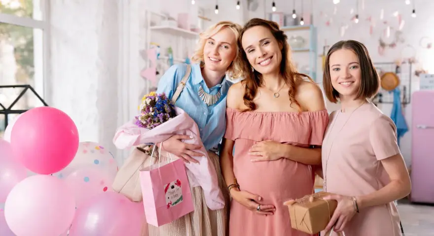 Trzy uśmiechnięte kobiety świętują baby shower. Jedna z nich jest w ciąży, a pozostałe dwie trzymają prezenty i balony.