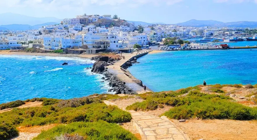 A stone path leads to a causeway connecting to the whitewashed town of Naxos, Greece, with vibrant blue waters lapping at the shore.