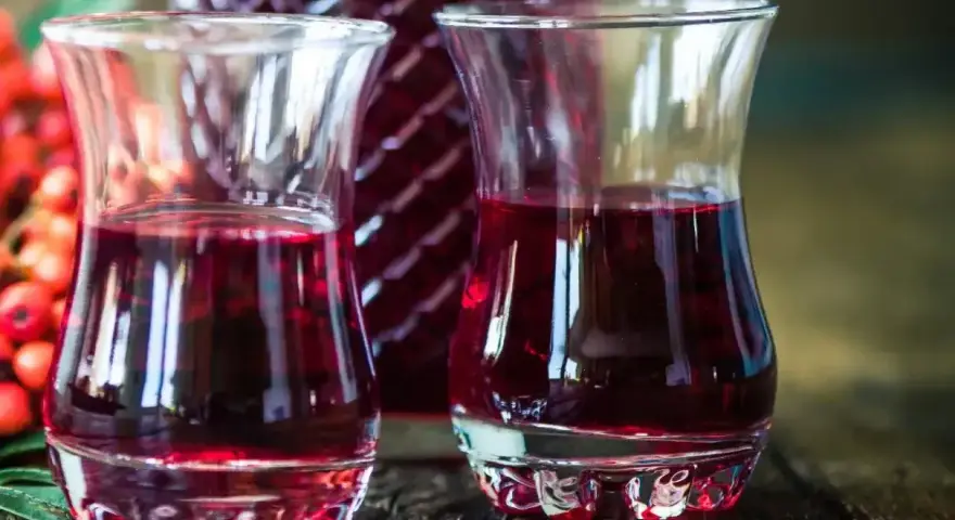 Dwa kieliszki wypełnione rubinową nalewką z czeremchy, obok butelka i kiść owoców.
