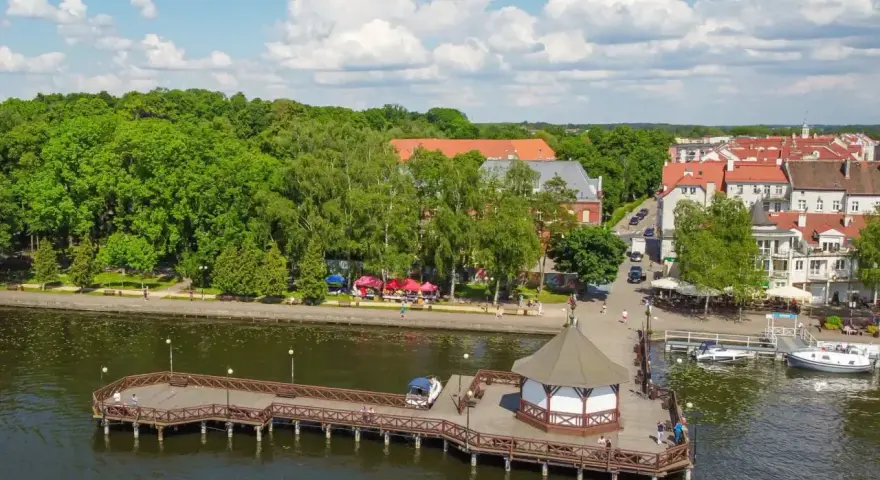 Malownicze mazury miejscowości nad jeziorem. Drewniane molo z altaną, zielone drzewa i urokliwe kamienice tworzą letni klimat.