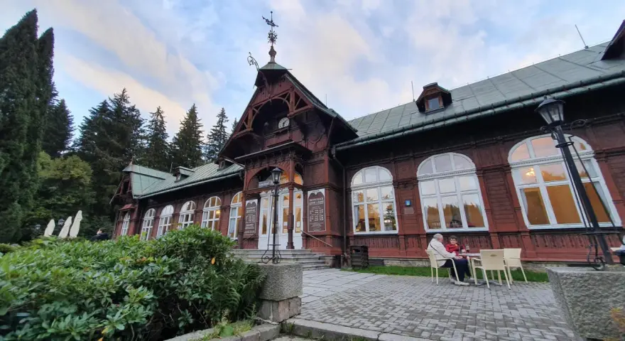 Drewniany pawilon z zegarem i karłowata studzienka przed wejściem. Na zewnątrz siedzą ludzie przy stoliku.