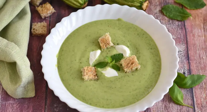 Crema fredda di zucchine servita in una ciotola bianca, guarnita con crostini, panna e menta. Fresca e invitante.