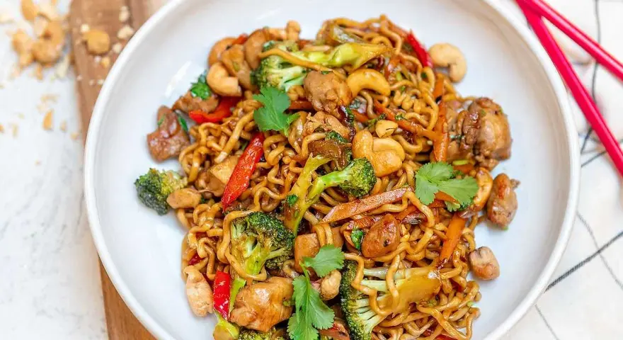 En skål med nudlar, kyckling, broccoli, paprika och cashewnötter. En perfekt allt i ett mat-rätt, redo att ätas med röda ätpinnar.
