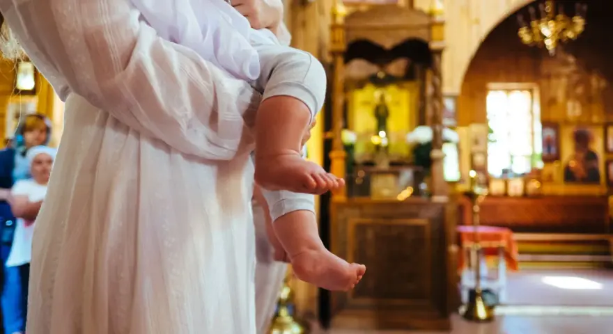 Chrzestna trzyma dziecko w kościele. W tle widać ikonostas i ławki.