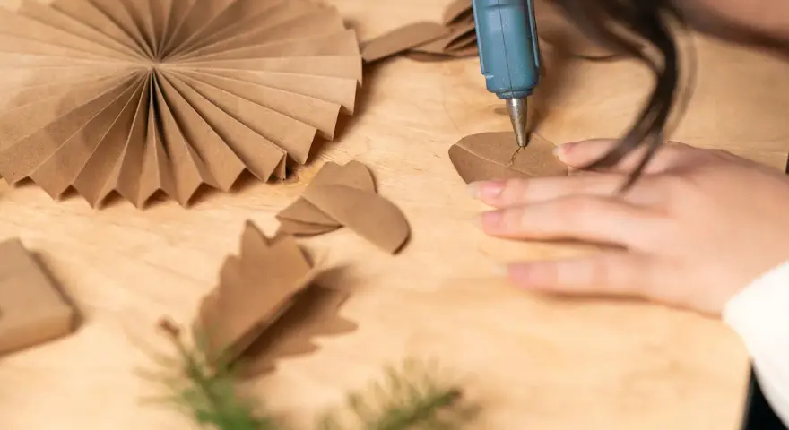 Manos creando manualidades con cartón para adultos. Se usa una pistola de silicona para pegar piezas de cartón, formando adornos.