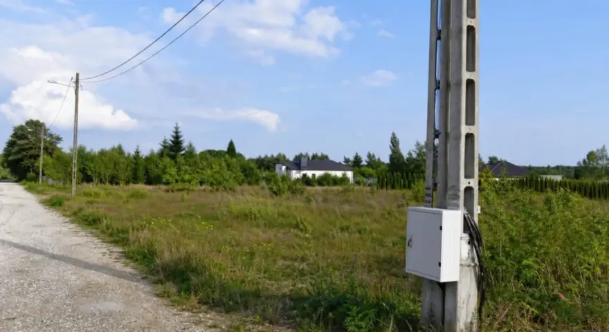 Betonowe słupy energetyczne stoją na tle zieleni i nieba. W oddali widać domki jednorodzinne.