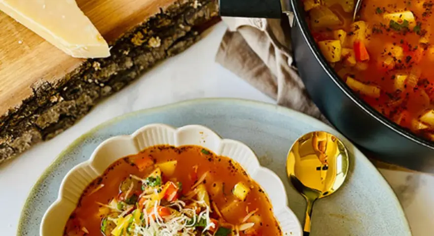 Aksamitna zupa minestrone z kawałkami warzyw i startym parmezanem, podana w ozdobnej misce. Obok garnek z zupą i kawałek sera na desce.