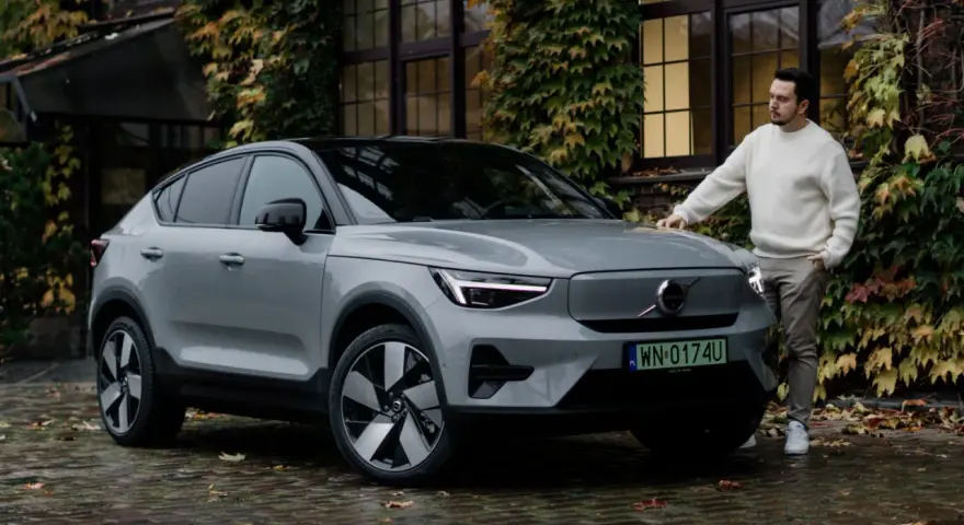 Szary Volvo C40 z czarnym dachem stoi na mokrej, brukowanej nawierzchni. Mężczyzna w białym swetrze opiera się o maskę.