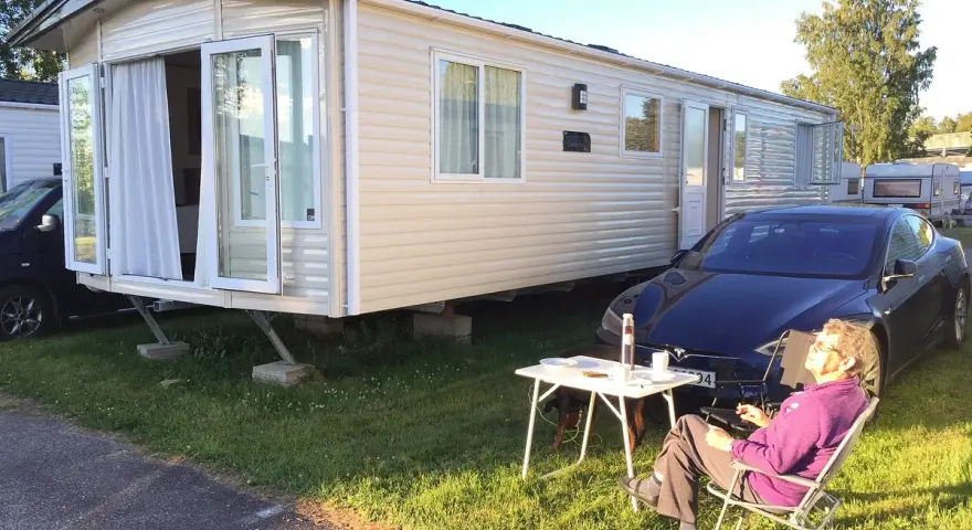 En person njuter av solen vid ett campingbord utanför en husvagn. En Tesla parkerad bredvid. Camping Timrå.