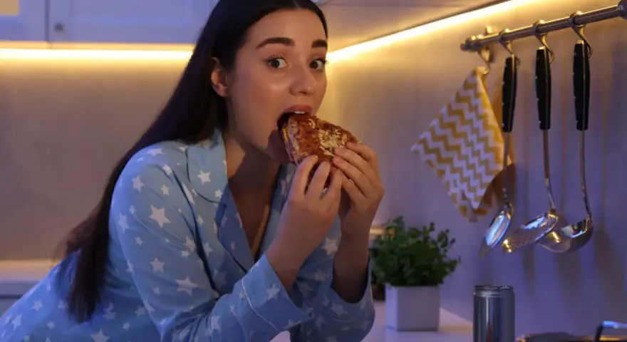 Kobieta w piżamie z gwiazdkami je kanapkę, choć czuje, że jest głodna, ale nie ma apetytu. Na blacie leży też pizza i burger.