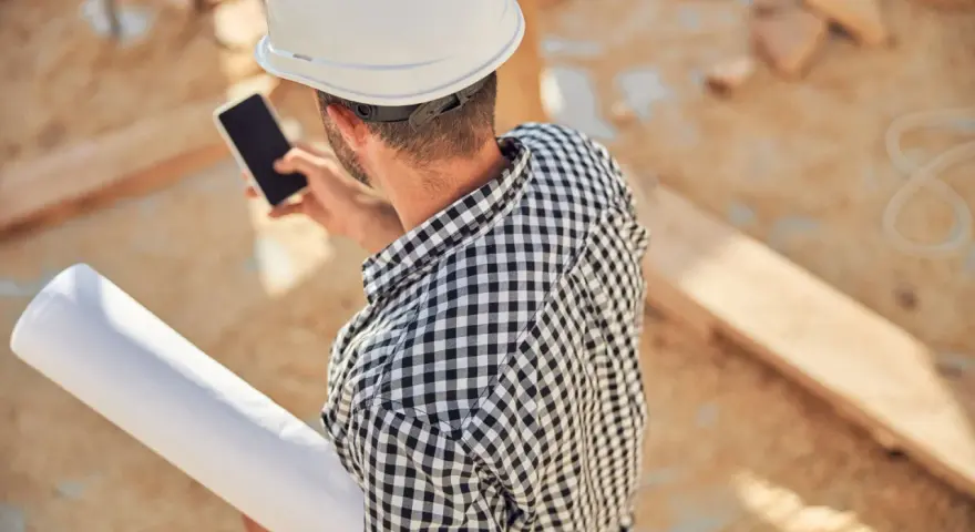 Budowlaniec w kasku sprawdza na telefonie dane o nośności gruntu kpa, trzymając zwój planów.