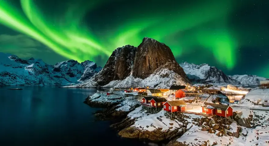 Grön norrsken dansar över snöklädda berg och röda fiskestugor vid en vik i Norge.