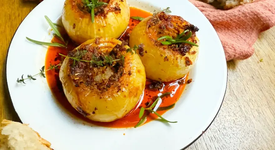 Tres cebollas al horno, rellenas y doradas, nadan en una salsa roja vibrante. Hierbas frescas coronan cada una.
