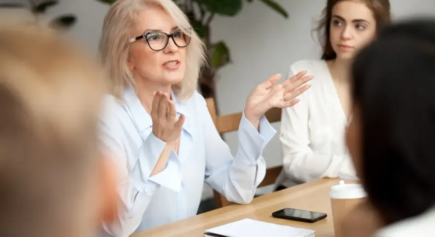 Kobieta w okularach gestykuluje, prowadząc dyskusję podczas rady pedagogicznej.