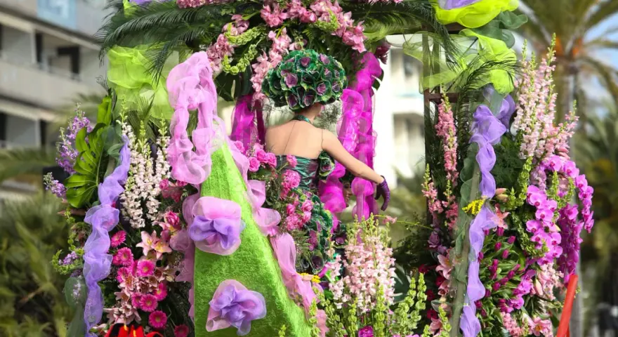 Una figura con un vestido floral y un tocado verde, rodeada de exuberantes flores rosas y moradas, participa en el **carnaval de Nice**.