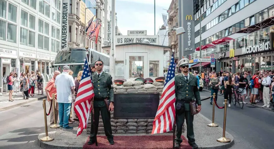 Zwei Männer in Uniformen mit US-Flaggen stehen am Checkpoint Charlie in Berlin. Touristen und ein McDonald's im Hintergrund.