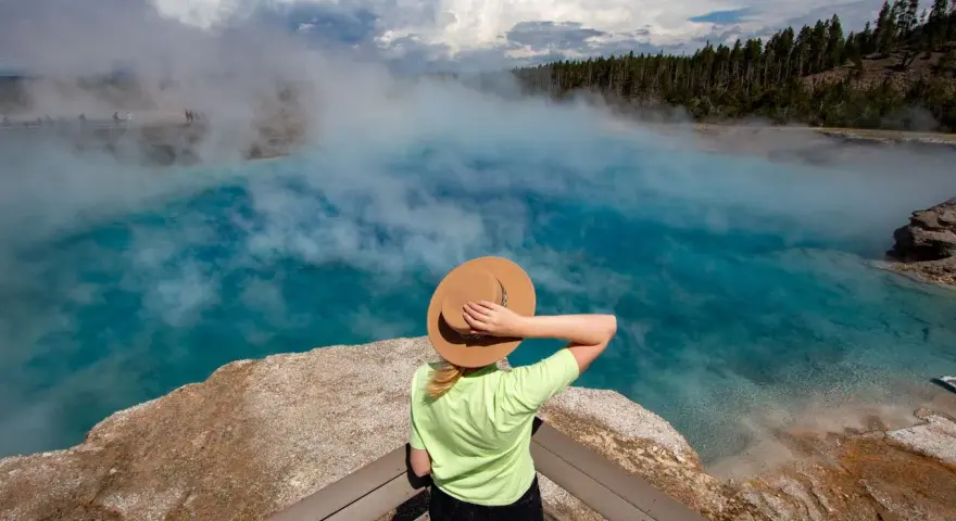Kobieta w kapeluszu podziwia turkusowe gorące źródło w Yellowstone. Gdzie jest Yellowstone? Tu, wśród parujących wód i lasów.