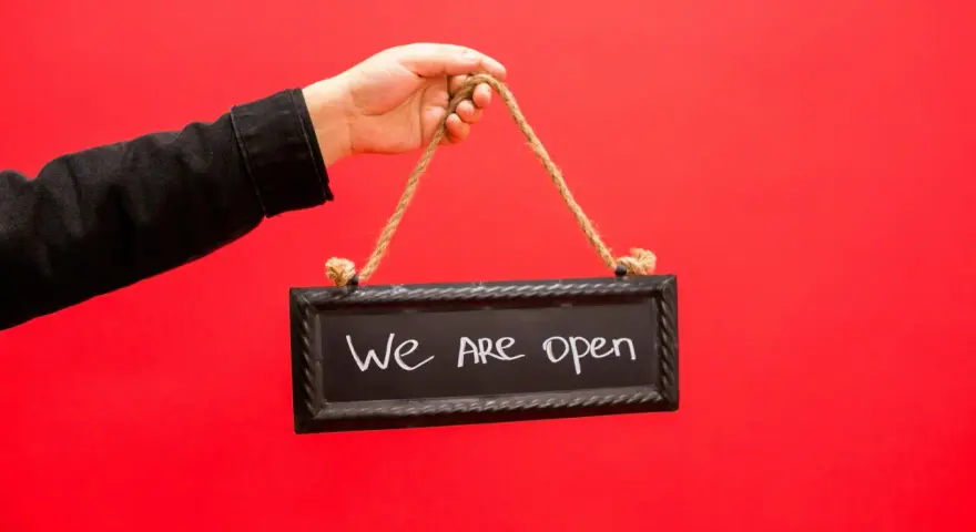 Mano con chaqueta negra sostiene un cartel que dice "We are open", indicando la apertura de un negocio, similar a los tipos de licencia de actividad.