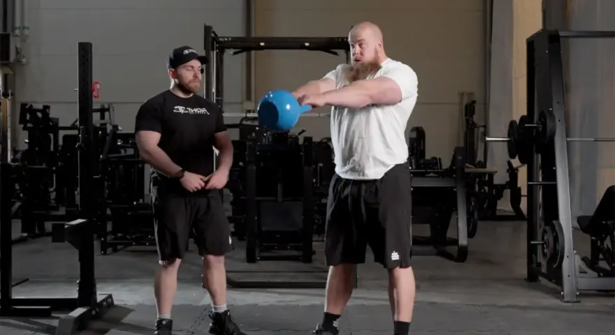 Man med skägg utför kettlebell swing varje dag i gym. Bredvid står en tränare.