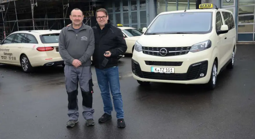 Zwei Männer stehen vor einem gelben Taxi und einem Kombi. Die Taxifahrt und der Steuersatz sind wichtige Themen für sie.