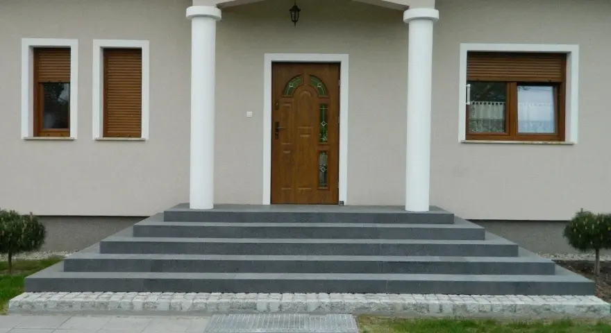 Eleganckie, szare schody zewnętrzne prowadzące do drewnianych drzwi. Świetny pomysł na schody zewnętrzne, podkreślający nowoczesny styl domu.