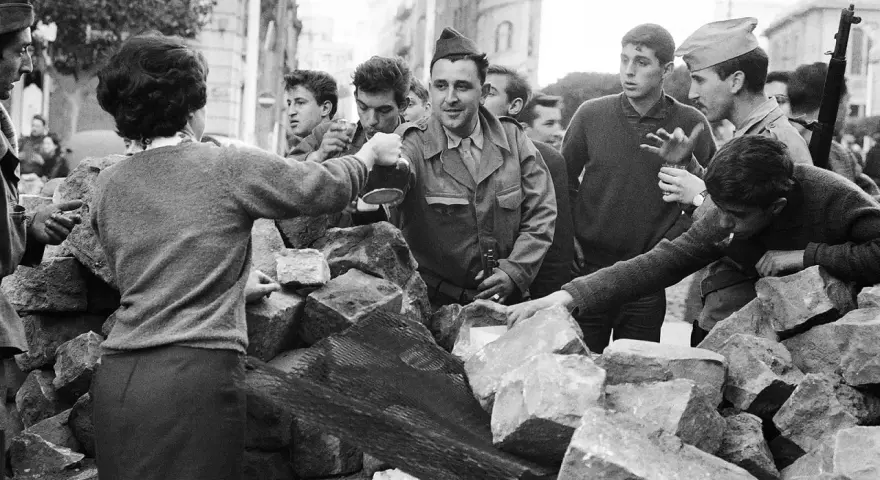 Un grupo de jóvenes y soldados interactúan tras una barricada de piedras en Argelia. Un hombre con uniforme militar sirve bebida.