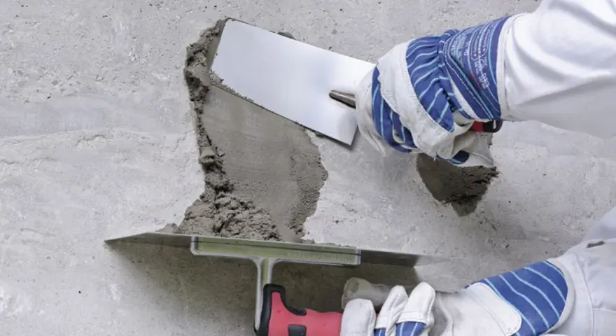 Naprawa betonu na zewnątrz. Ręce w rękawicach nakładają zaprawę kielnią na uszkodzony fragment ściany.