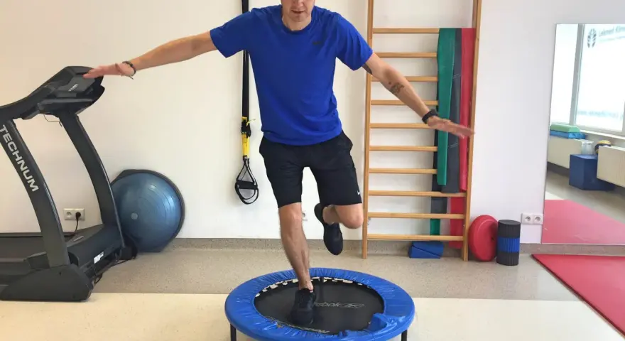 Mężczyzna wykonuje ćwiczenia po rekonstrukcji ACL, balansując na małej trampolinie. W tle bieżnia i drabinka gimnastyczna.