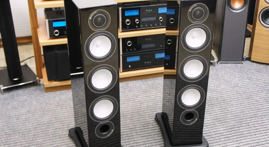 Dwie eleganckie kolumny głośnikowe Monitor Audio RX8 w czarnym połysku, stojące na szarym dywanie, obok sprzętu audio.