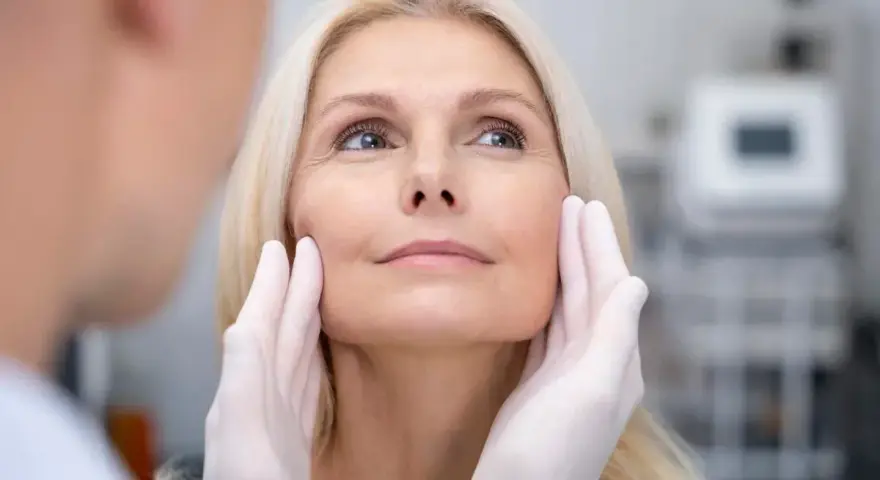 Kobieta z blond włosami poddaje się zabiegowi depilacji laserowej twarzy. Dłonie w rękawiczkach delikatnie dotykają jej policzków.