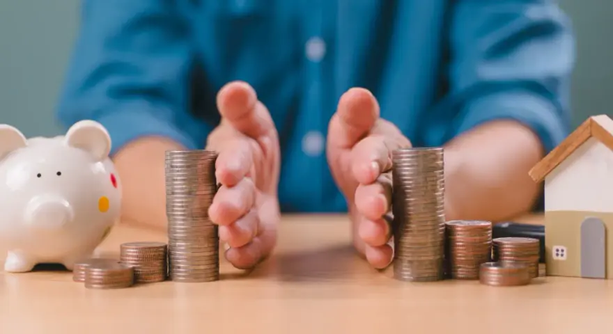 Mains protégeant des piles de pièces, symbolisant la gestion financière et la **desolidarisation pret immo**. Une tirelire et une maison miniature sont visibles.