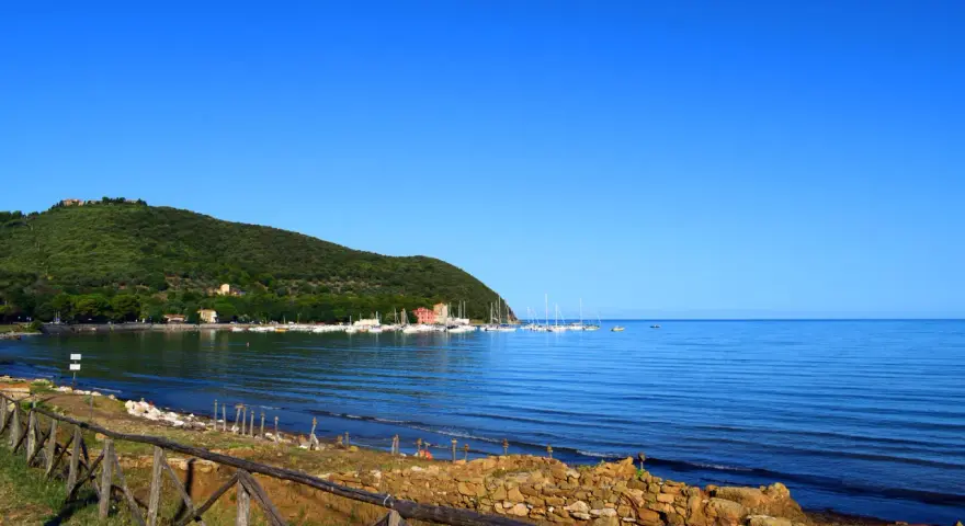 A tranquil bay in Tuscany with sailboats docked, a green hillside, and a rustic wooden fence in the foreground.