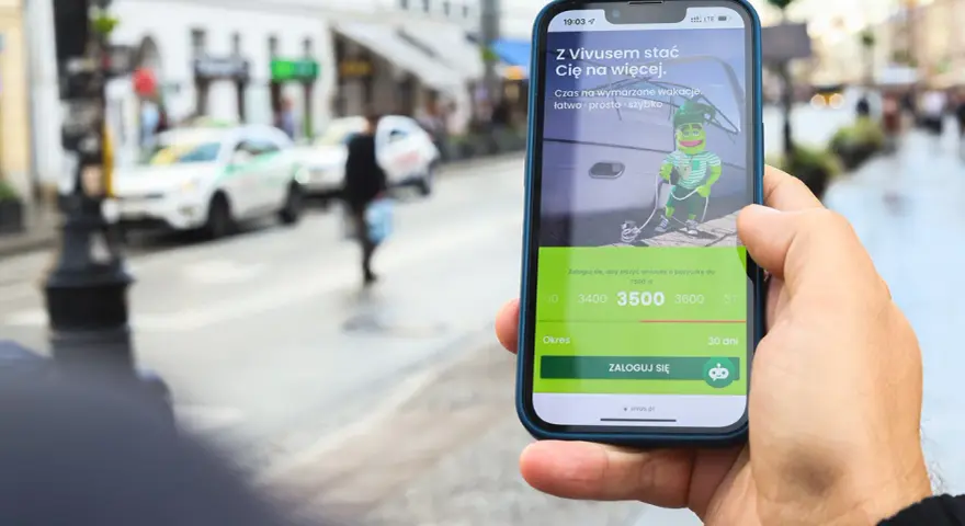 Dłoń trzyma telefon z aplikacją Vivus, gdzie można złożyć wniosek o pożyczkę. W tle miejski krajobraz. Vivus windykacja pomaga spełniać marzenia.