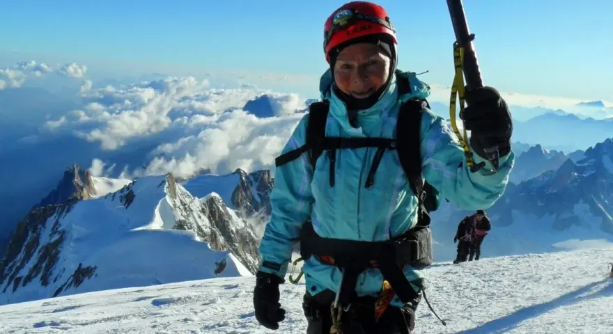 Barbara Prymakowska, mimo swojego wieku, zdobywa szczyty. Uśmiechnięta alpinistka w kasku i turkusowej kurtce na tle gór i chmur.