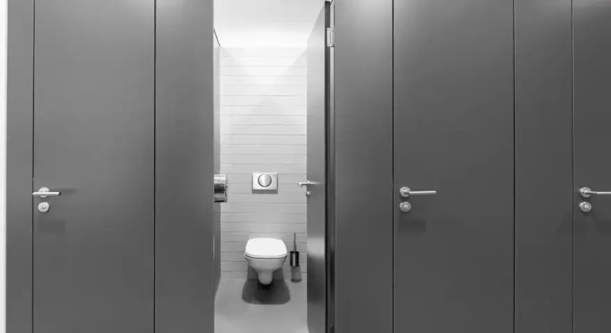Reihe von grauen Türen zu Toilettenkabinen. Eine Tür ist geöffnet und gibt den Blick auf eine Toilette frei. Gute Frage, wie viele Toiletten pro Mitarbeiter?
