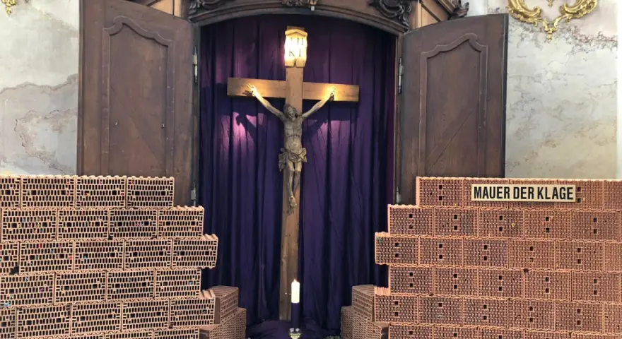 Kreuzweg Stationen: Jesus am Kreuz, umgeben von Ziegelmauern, die die "Mauer der Klage" symbolisieren. Eine Kerze brennt vor dem Kruzifix.
