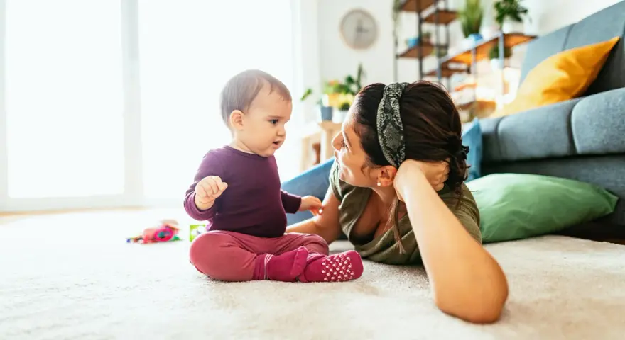 Mama patrzy na dziecko, które siedzi na dywanie. To obrazek pokazujący, co powinien umieć 2 latek: rozwijać się i bawić.