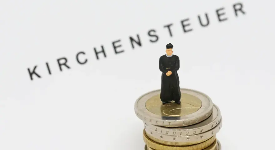 Miniatur-Priester steht auf Münzstapel. Der Schriftzug "Kirchensteuer" im Hintergrund wirft die Frage auf, warum man kirchensteuer zahlt.