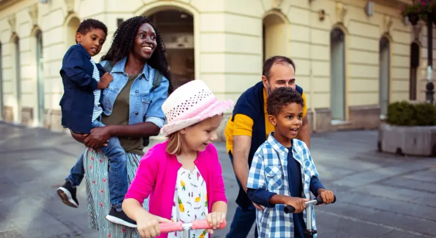 Glückliche Familie unterwegs: Mutter trägt Kind, Vater schiebt Roller. Ein Spagat zwischen Kind und neuem Partner gelingt hier spielerisch.