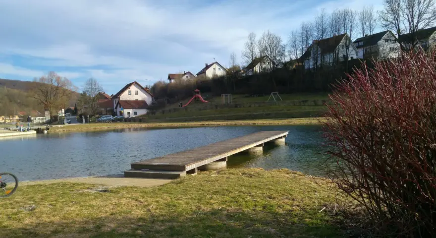 Holzsteg am Badesee Forchheim, mit Häusern im Hintergrund und einem Spielplatz.