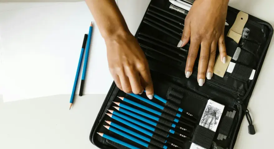 Un étui ouvert révèle une sélection de crayons de différents types pour dessiner, avec des mains choisissant un crayon bleu.