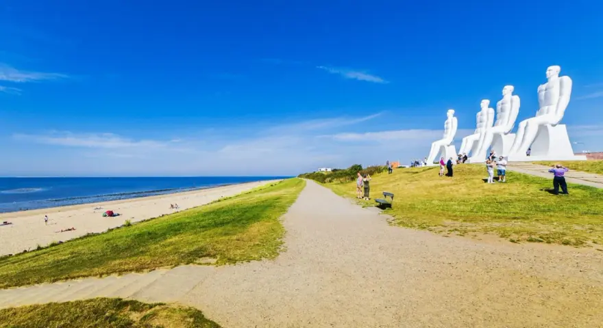 De vita sandstränderna i Danmark möter en grönskande sluttning med gigantiska sittande figurer. En grusväg leder mot havet under en klarblå himmel.