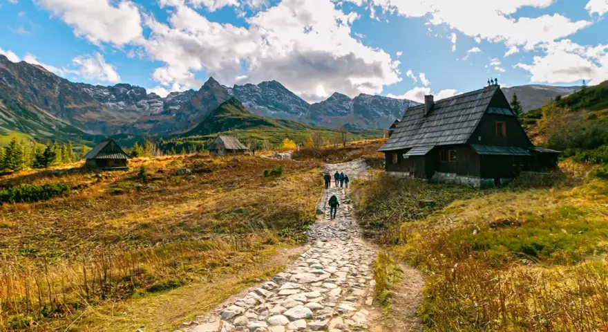Kamienista ścieżka prowadzi do Rusinowej Polany, gdzie widać drewniane chaty i turystów. W tle majestatyczne Tatry.