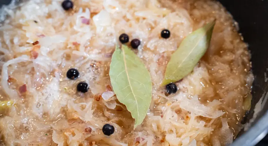 Sauerkraut köchelt mit Lorbeerblättern und Wacholderbeeren, um den Geschmack zu verfeinern. Kleine rote Zwiebelstücke sind ebenfalls sichtbar.