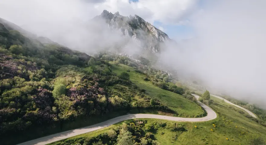 Strada tortuosa che sale verso le vette nebbiose, evocando l'ascesa al **Col du Tourmalet**.