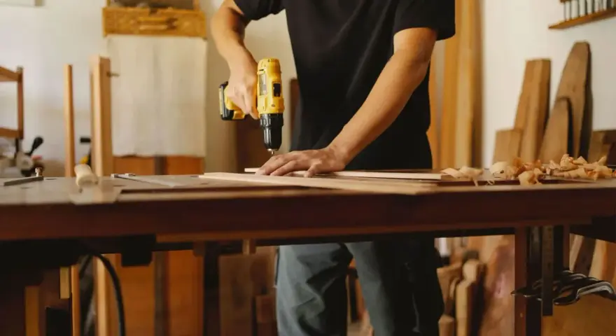 Mann bohrt Holzbretter, um seine Werkstatt einzurichten. Hobelspäne liegen auf dem Tisch.