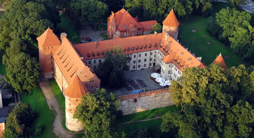Zamek w Bytowie otoczony zielenią, z czerwonymi dachami i okrągłymi wieżami. Na dziedzińcu widać stoliki i parasole.
