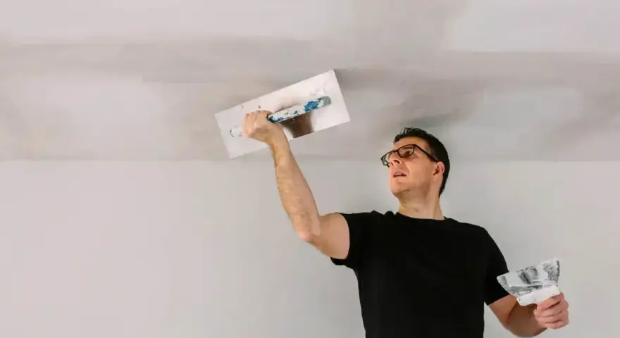Mężczyzna w okularach wykonuje tynkowanie sufitu, używając kielni i packi.