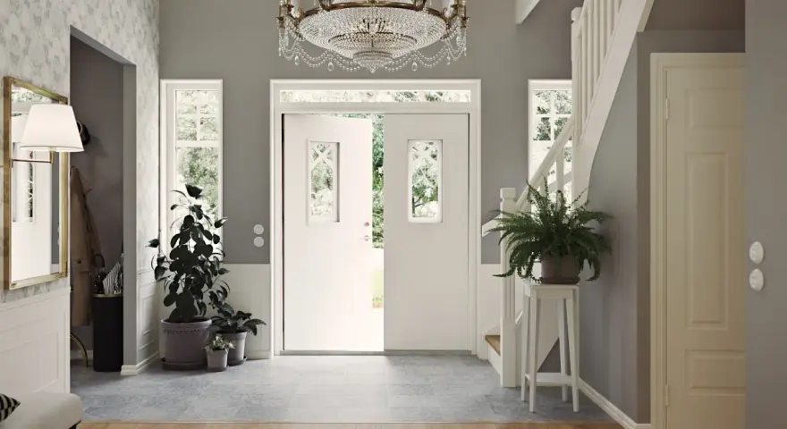Välkomnande hall med dubbeldörrar, kristallkrona och trappa. Gröna växter ger liv åt det inredda utrymmet.
